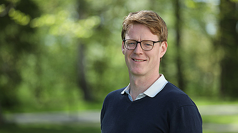 Mann mit Brille und dunkelblauem Pullover vor unscharf grünem Hintergrund.